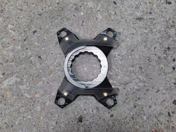 A black metal four-spoke wheel spacer with a machined silver center ring, photographed from above on a gray concrete surface.