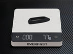 A white digital pocket scale with a black display screen and a black handle on top, resting on a black carbon fiber textured surface. The brand name 'OVERFAST' is clearly visible on the front.