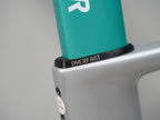 Close-up of a teal bicycle seat clamp with the brand name 'OVERFAST' printed in white on a black ring, attached to a silver frame against a neutral gray background.