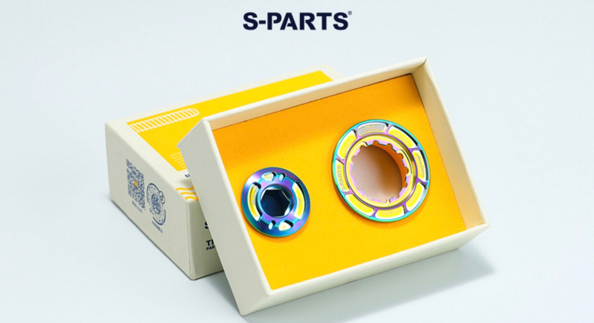 Two metallic mechanical parts displayed inside an open cream-colored box with a yellow insert, alongside another closed box. The brand name 'S-PARTS' is printed in dark blue at the top of the image.