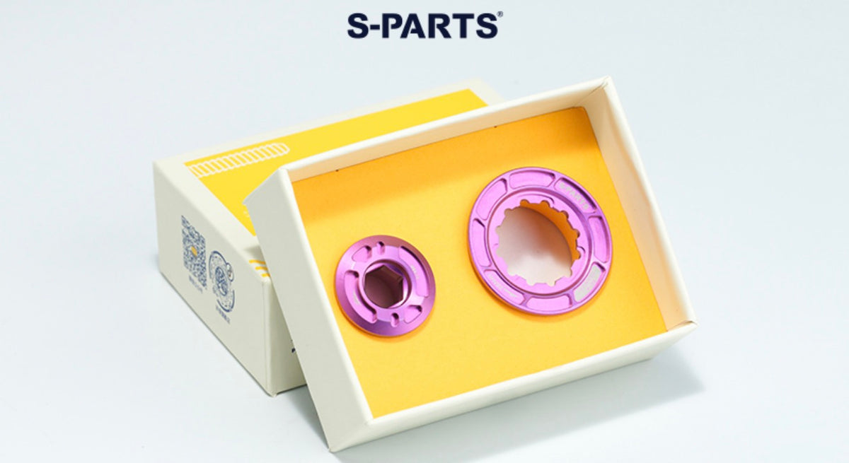 Two metallic purple mechanical parts displayed inside an open white box with a yellow insert, with the S-PARTS logo above on a light gray background.