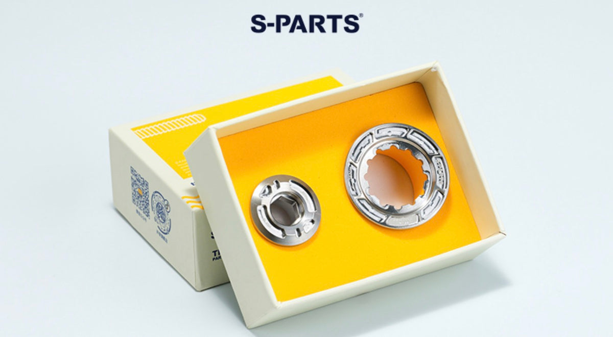 Two metallic mechanical parts displayed inside an open cream-colored box with a yellow insert, alongside a branded S-PARTS box on a clean white background.