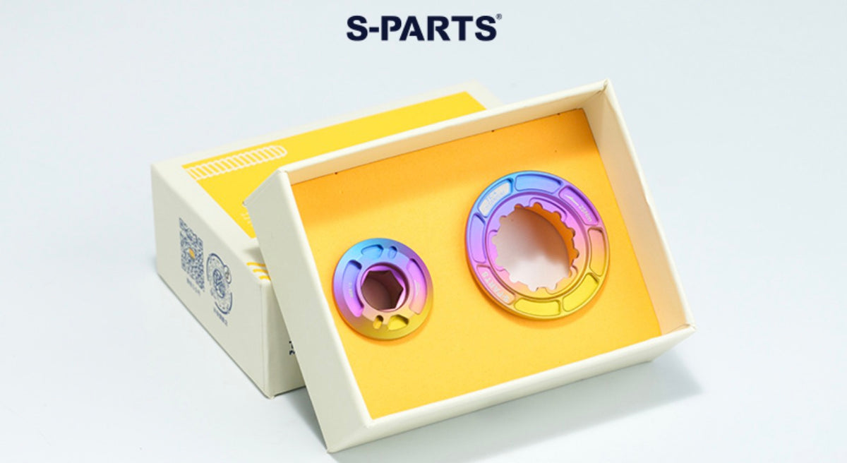Two circular metallic parts with an iridescent rainbow finish displayed inside an open cream-colored box with a yellow insert. The brand name 'S-PARTS' is printed in black at the top of the image.